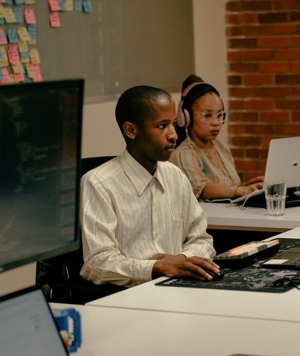 Three professionals working on computers in a modern office with a brick wall and a bulletin board with colorful sticky notes.